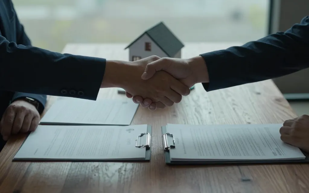 Two people shaking hands across a table with a signed contract and house model between them.